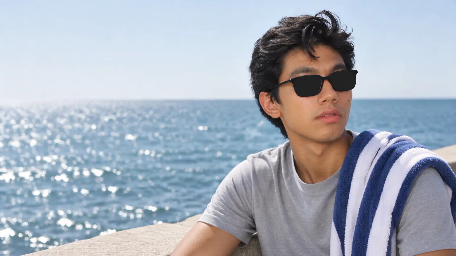 A young man in black sunglasses with solid black lenses sits by a bright, glittering sea, a striped towel over his shoulder.